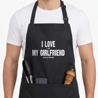 Play video game apron - White stacked text reading I love it when my girlfriend lets me play video games on black fabric — shown on a black cotton canvas apron against a white background
