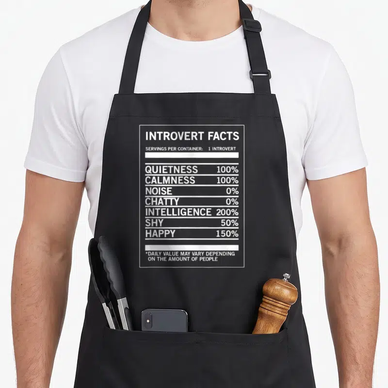 Introvert witty aprons - White nutrition label style text reading Introvert Facts with percentages for quietness — shown on a black cotton canvas apron against a white background