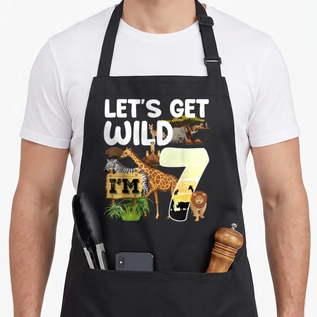 Animal 7th birthday apron - Giraffe zebra lion meerkats pangolin elephant cheetah vulture rhino birds with LET'S GET — shown on a black cotton canvas apron against a white background