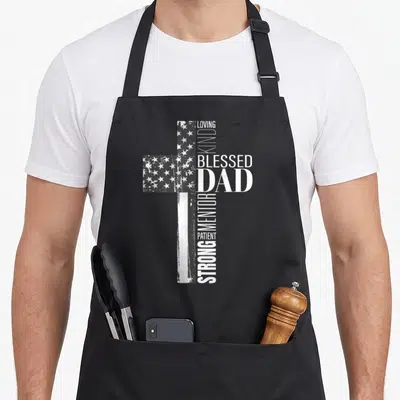 Christian american flag apron - Distressed cross with flag pattern and words LOVING KIND BLESSED DAD STRONG MENTOR — shown on a black cotton canvas apron against a white background