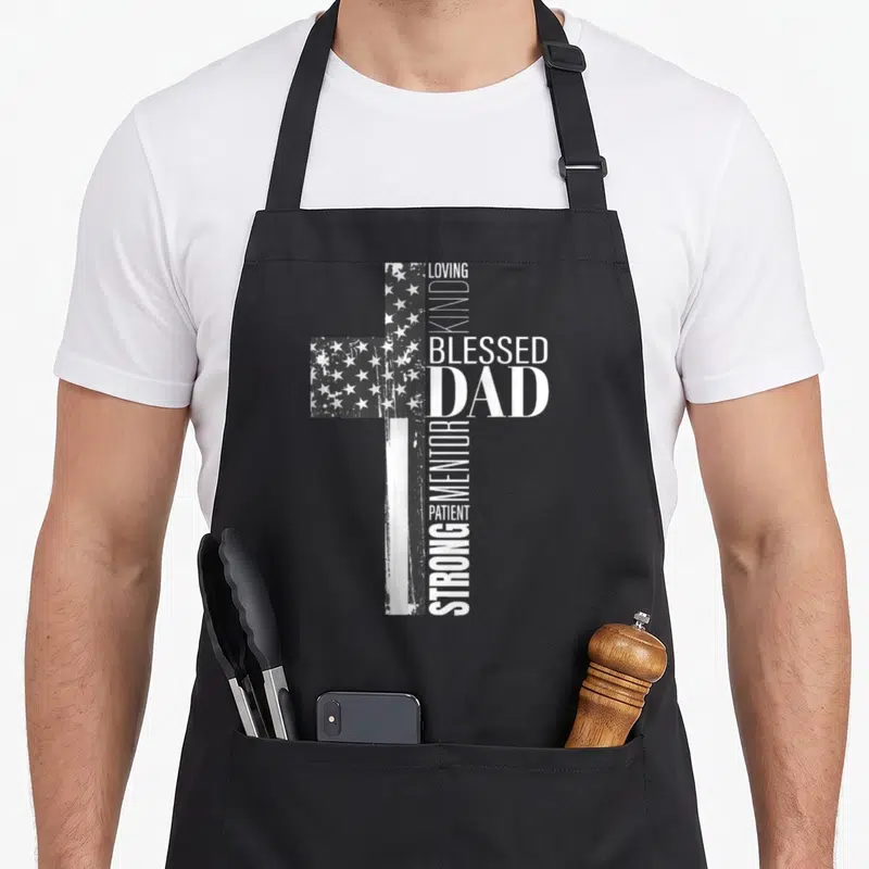 Christian american flag apron - Distressed cross with flag pattern and words LOVING KIND BLESSED DAD STRONG MENTOR — shown on a black cotton canvas apron against a white background