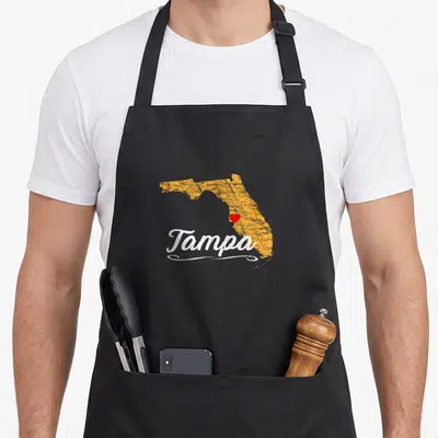 Tampa bay florida apron - Distressed orange Florida map with white Tampa text and red heart marker over city location — shown on a black cotton canvas apron against a white background