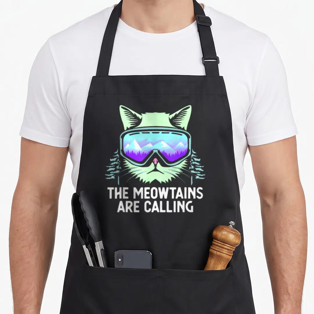 Snowboard winter apron - Cat head with ski goggles reflecting blue purple mountains and mint green pine trees — shown on a black cotton canvas apron against a white background