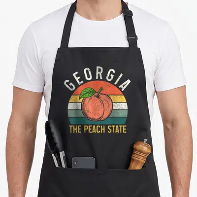Georgia peach pride apron - Retro striped sun with peach and GEORGIA THE PEACH STATE text — shown on a black cotton canvas apron against a white background