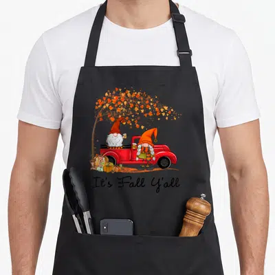 Yall fall aprons - Two gnomes in red truck under autumn tree with It's Fall Y'all text below — shown on a black cotton canvas apron against a white background