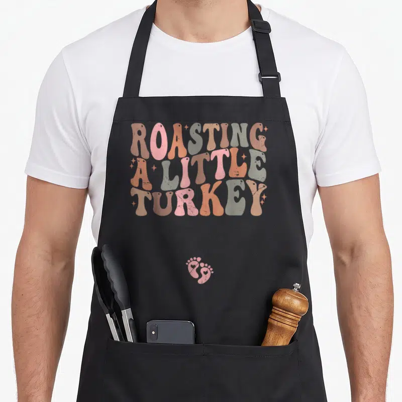 Roasting thanksgiving apron - Retro ROASTING A LITTLE TURKEY text in pink orange tan with two baby footprints below — shown on a black cotton canvas apron against a white background