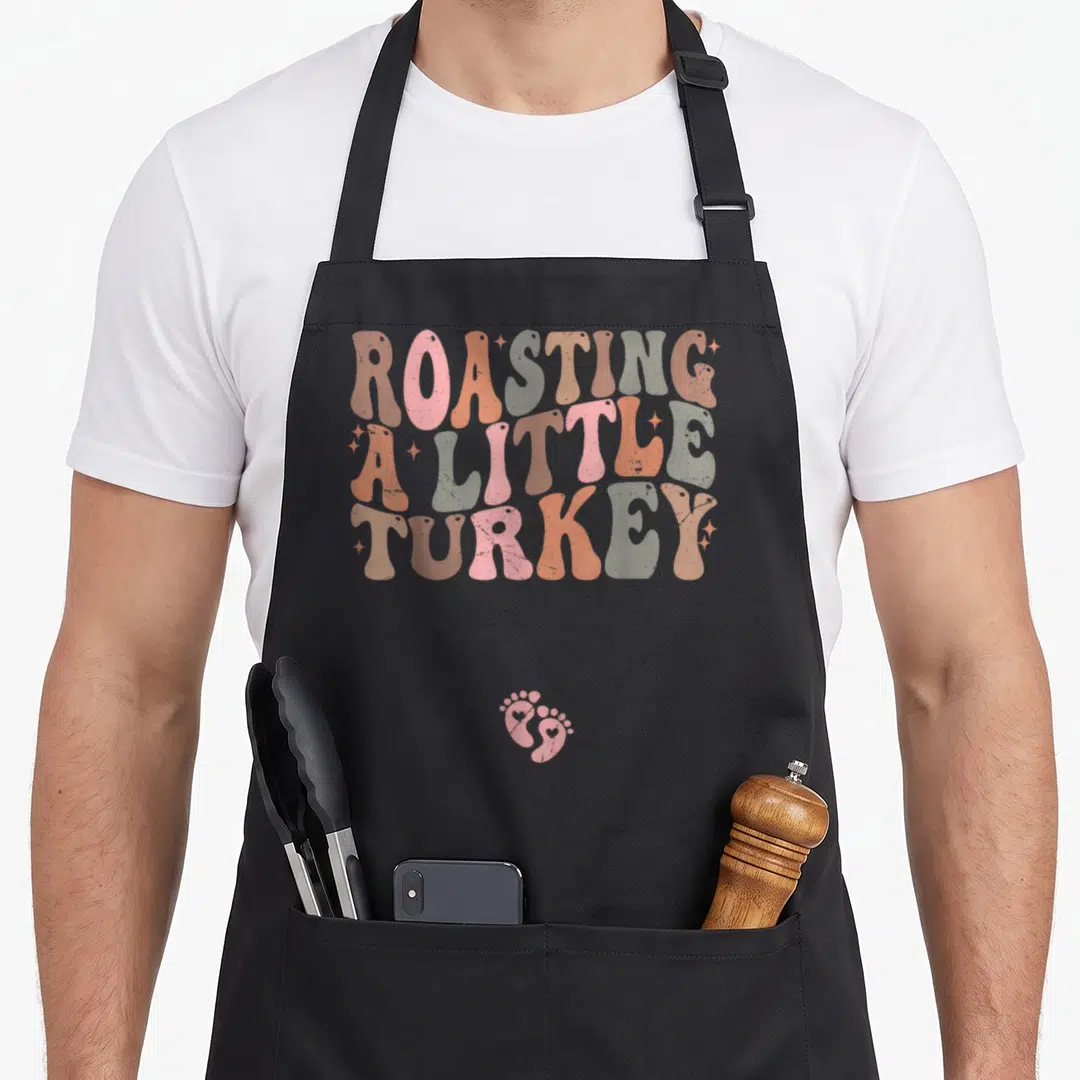 Roasting thanksgiving apron - Retro ROASTING A LITTLE TURKEY text in pink orange tan with two baby footprints below — shown on a black cotton canvas apron against a white background