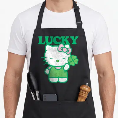 Clover lucky apron - Hello Kitty standing holding four-leaf clover under LUCKY text in green and pink — shown on a black cotton canvas apron against a white background