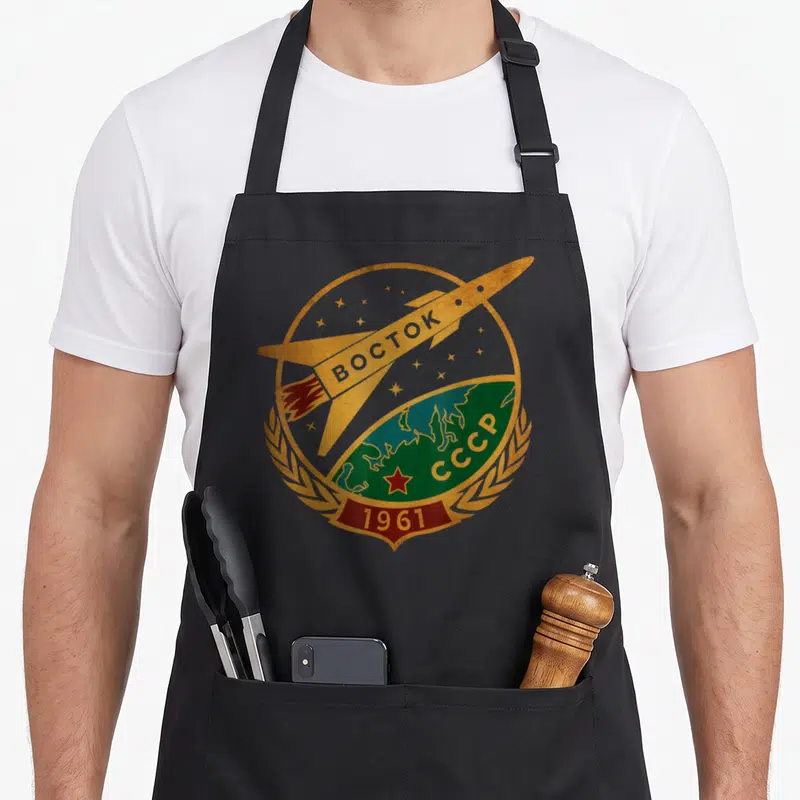 Check russia apron - Retro gold and red space mission badge with rocket and globe on black fabric — shown on a black cotton canvas apron against a white background