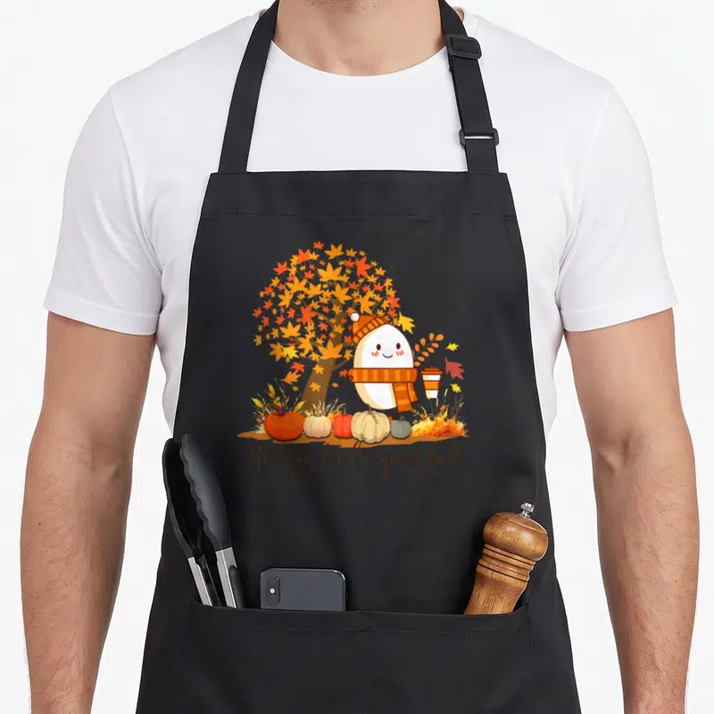 Dumpty thanksgiving cute fall aprons - Cartoon egg in scarf holding cup near pumpkins and autumn tree with text — shown on a black cotton canvas apron against a white background