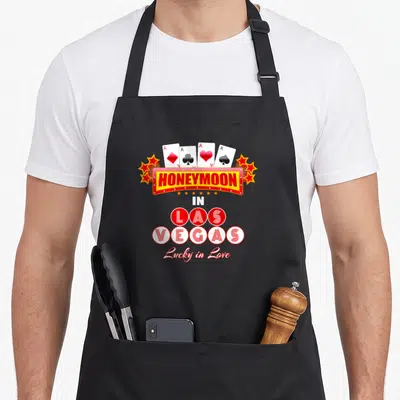 Las newlywed aprons - Four playing cards above Honeymoon in Las Vegas sign and Lucky in Love text — shown on a black cotton canvas apron against a white background
