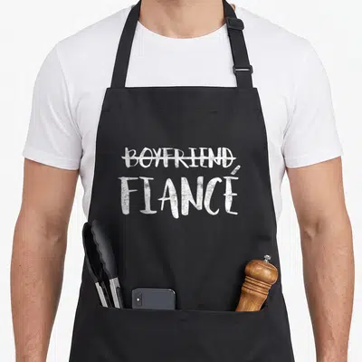 Engagement wedding apron - White text reading BOYFRIEND crossed out above FIANCÉ — shown on a black cotton canvas apron against a white background