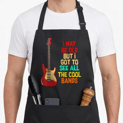 See but girl apron - Red electric guitar next to seven lines of multi-colored text reading i may be old but i got — shown on a black cotton canvas apron against a white background