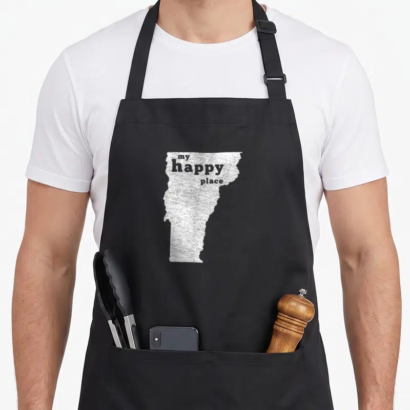 Distressed vermont apron - White Vermont state silhouette containing the text my happy place — shown on a black cotton canvas apron against a white background
