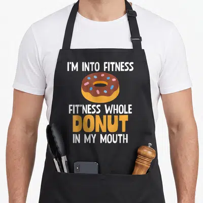 Fitness donut apron - Cartoon donut with brown icing and sprinkles surrounded by white and orange text — shown on a black cotton canvas apron against a white background