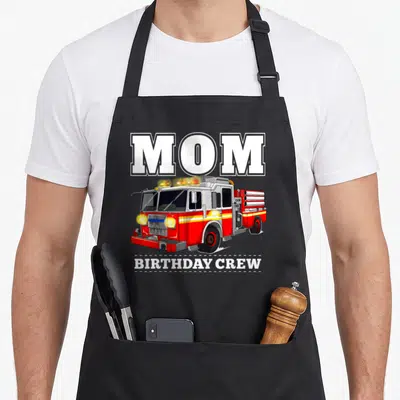 Fire crew aprons - MOM BIRTHDAY CREW text above red white and yellow fire truck illustration — shown on a black cotton canvas apron against a white background