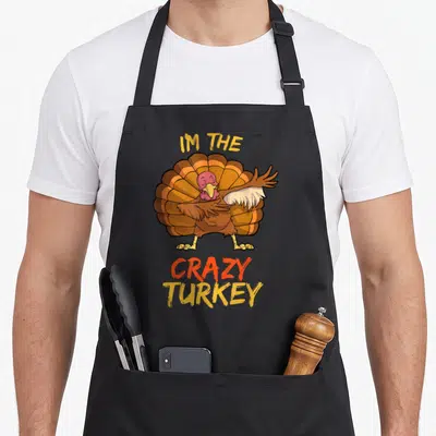 Cute family thanksgiving apron - Cartoon turkey with spread wings and IM THE CRAZY TURKEY text in brown orange — shown on a black cotton canvas apron against a white background