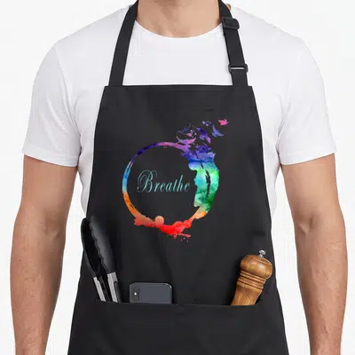 Heavenly breathwork apron - Watercolor circle in purple blue green and red with Breathe text in light blue script — shown on a black cotton canvas apron against a white background