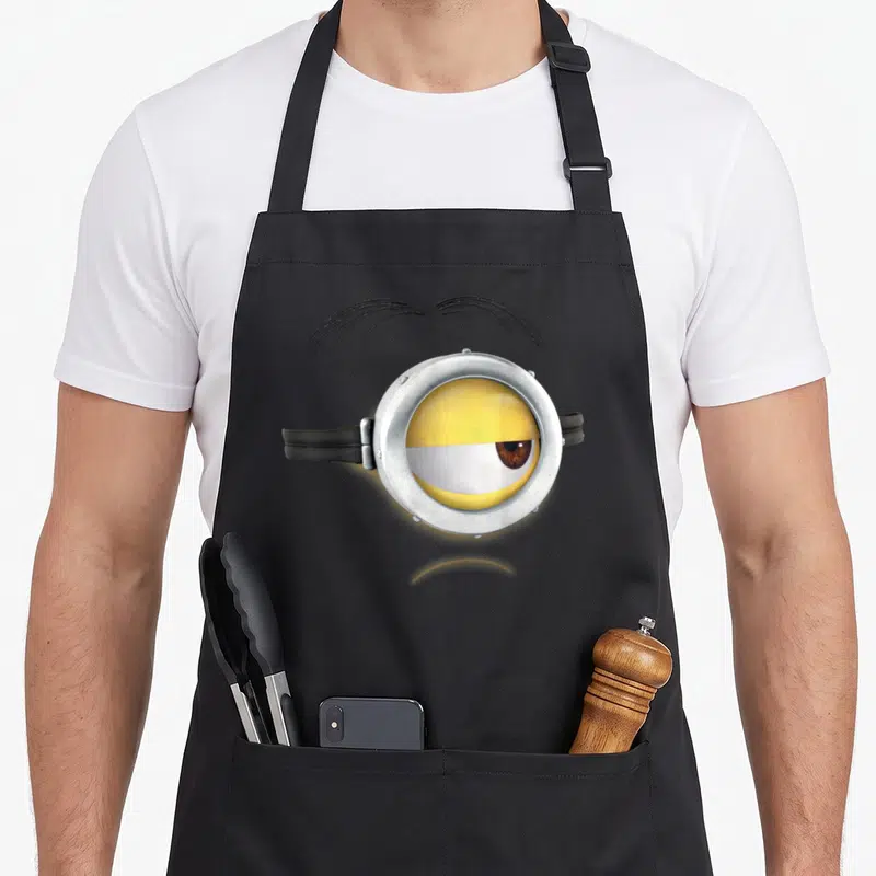 Frown face apron - Realistic yellow minion eye in silver goggle with black eyebrow and strap — shown on a black cotton canvas apron against a white background