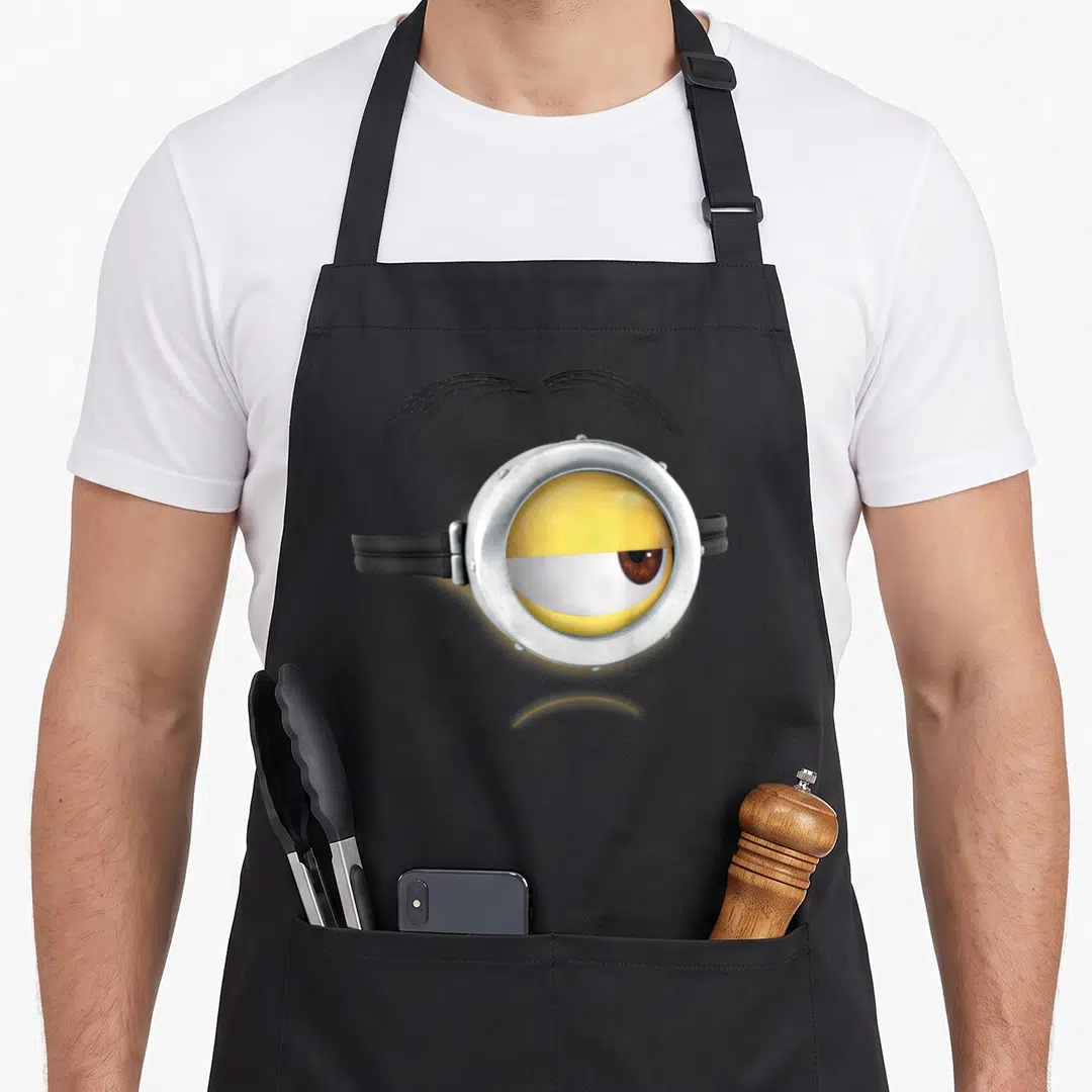 Frown face apron - Realistic yellow minion eye in silver goggle with black eyebrow and strap — shown on a black cotton canvas apron against a white background