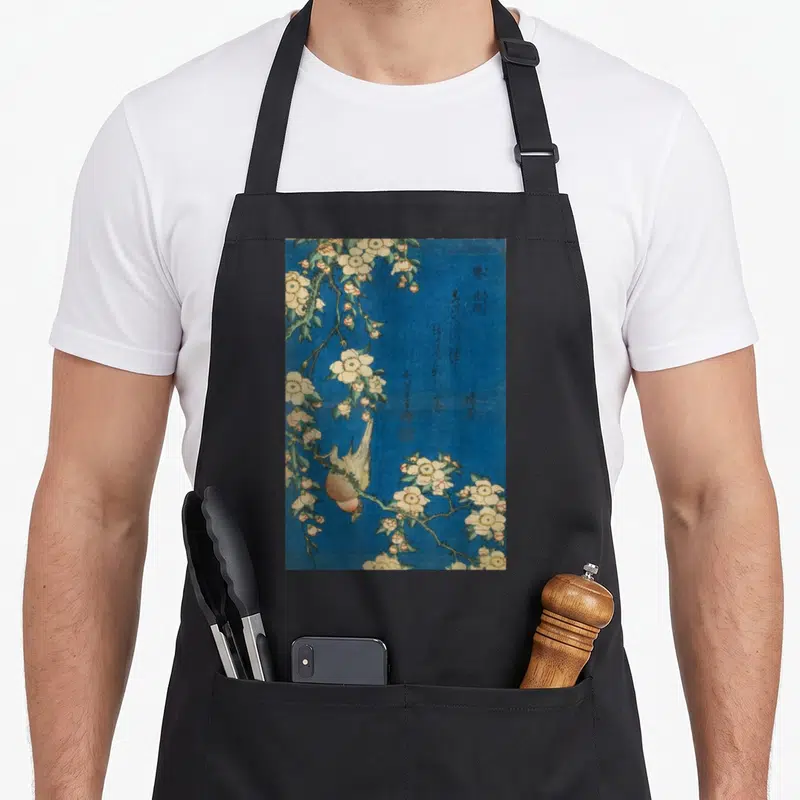 Goldfinch cherry apron - Realistic bird perched upside down on cherry blossoms with calligraphy on blue background — shown on a black cotton canvas apron against a white background