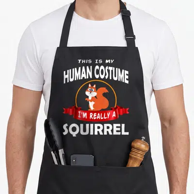 Human squirrel aprons - Cartoon squirrel in orange circle with red ribbon and text THIS IS MY HUMAN COSTUME I'M REALLY — shown on a black cotton canvas apron against a white background