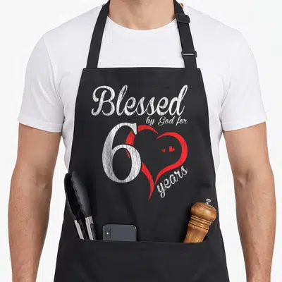 Blessed 60th birthday apron - Distressed white text Blessed by God for 60 years over red heart graphic — shown on a black cotton canvas apron against a white background