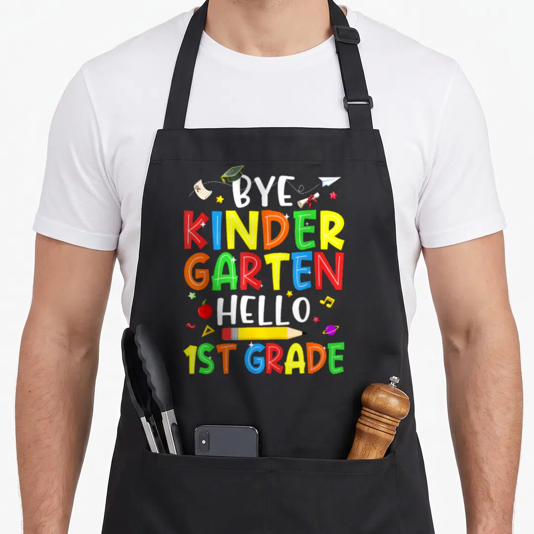 Graduation hello apron - Stacked text BYE KINDERGARTEN HELLO 1ST GRADE with pencil, apple, and cap icons in primary — shown on a black cotton canvas apron against a white background