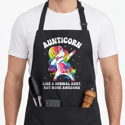 Normal unicorn apron - Dabbing rainbow unicorn with arched AUNTICORN text and scattered stars — shown on a black cotton canvas apron against a white background