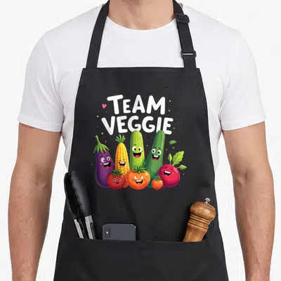 Playful veggie apron - Stacked Team Veggie text above cartoon eggplant, corn, cucumber, tomato, and beet with faces — shown on a black cotton canvas apron against a white background