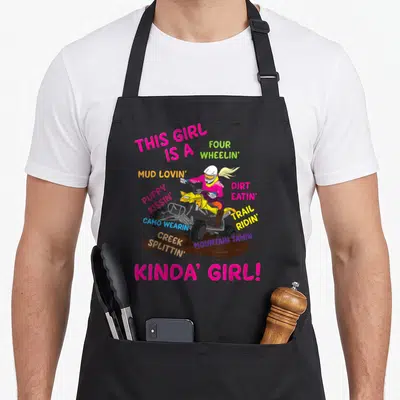 Atv fourth apron - Cartoon woman on yellow ATV in mud with pink and yellow arching text listing off-road traits — shown on a black cotton canvas apron against a white background