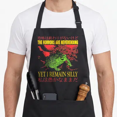 Horror frog aprons - Green toad illustration with red monster eyes and yellow English and red Japanese text — shown on a black cotton canvas apron against a white background