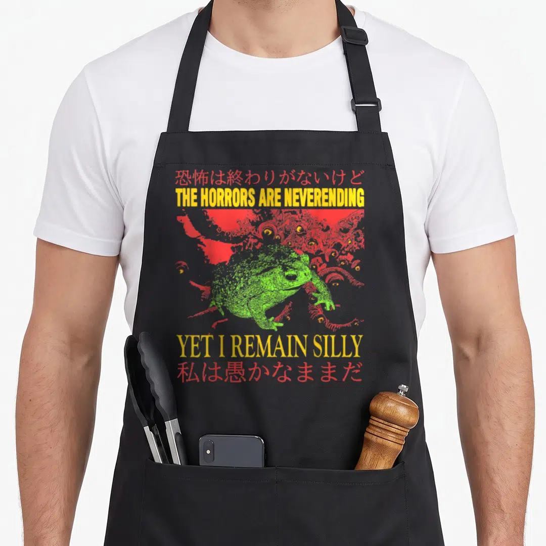 Horror frog aprons - Green toad illustration with red monster eyes and yellow English and red Japanese text — shown on a black cotton canvas apron against a white background