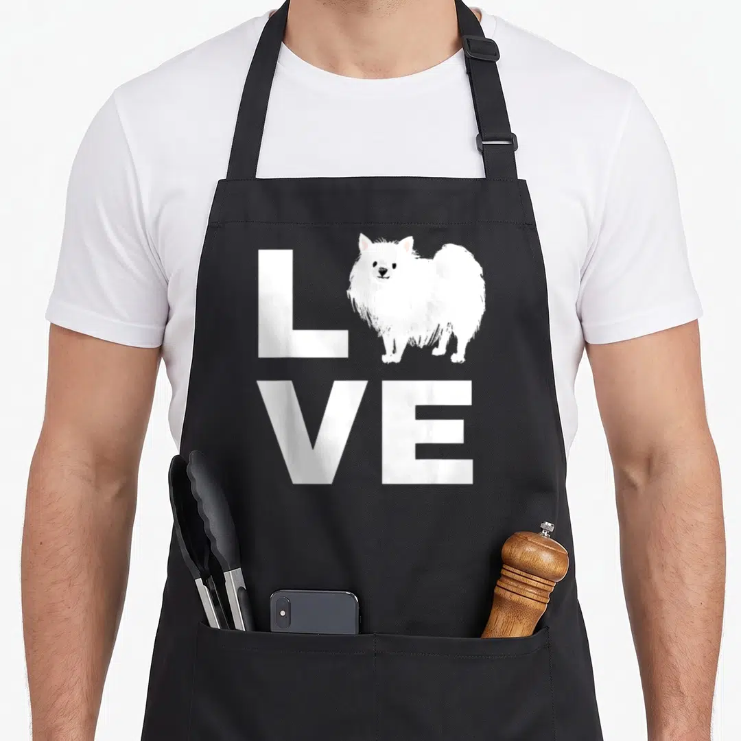 Pomeranian puppy dog apron - White cartoon Pomeranian dog standing in place of the letter O within the word LOVE — shown on a black cotton canvas apron against a white background