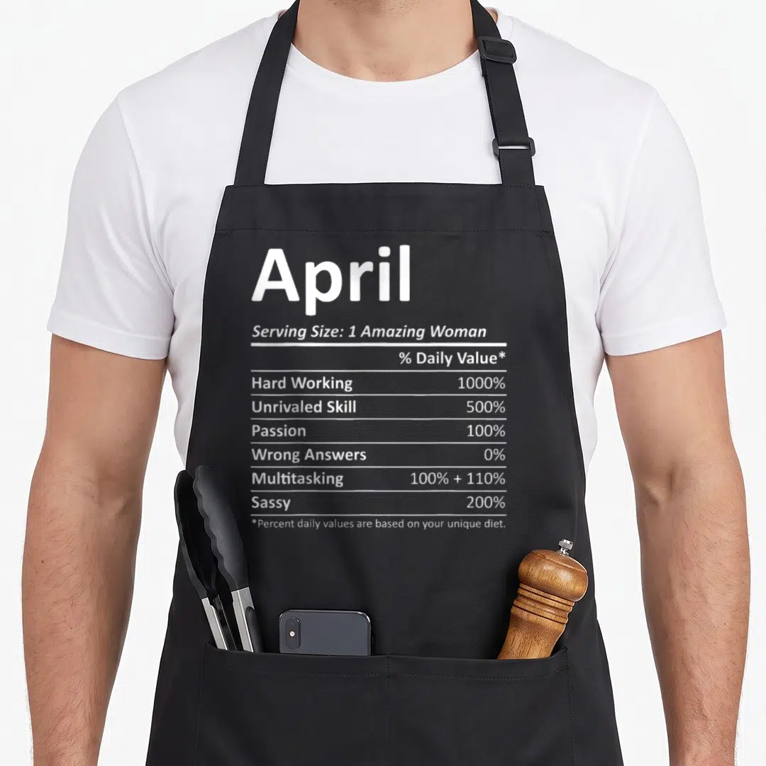 Nutrition april apron - April nutrition facts label with percentages for hard work, multitasking, and sass in white text — shown on a black cotton canvas apron against a white background