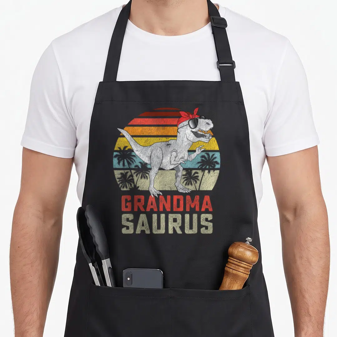 Grandmasaurus grandma apron - Retro T-Rex with sunglasses and red bandana, GRANDMA SAURUS text, striped sun background — shown on a black cotton canvas apron against a white background