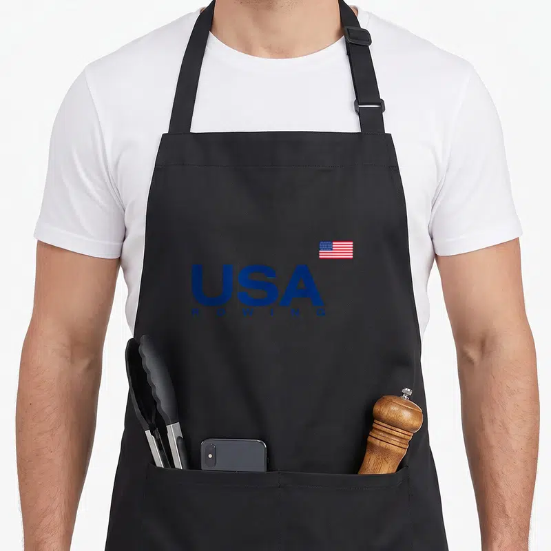 Flag rowing apron - Stacked blue USA ROWING text with a small American flag positioned above the right side — shown on a black cotton canvas apron against a white background