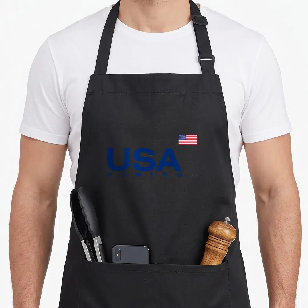 Flag rowing apron - Stacked blue USA ROWING text with a small American flag positioned above the right side — shown on a black cotton canvas apron against a white background