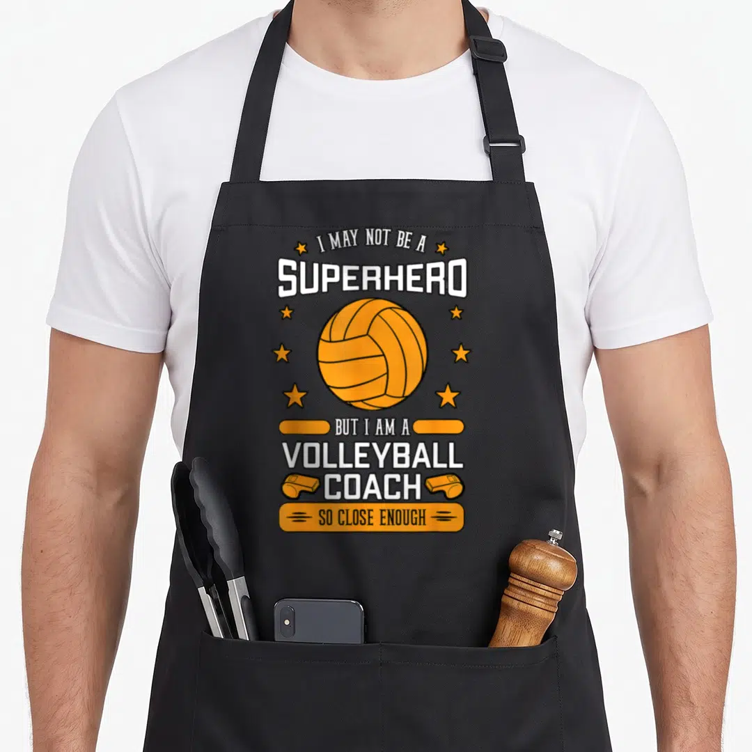 Superhero sports team aprons - Orange volleyball surrounded by stars and whistles with stacked text about being a coach — shown on a black cotton canvas apron against a white background