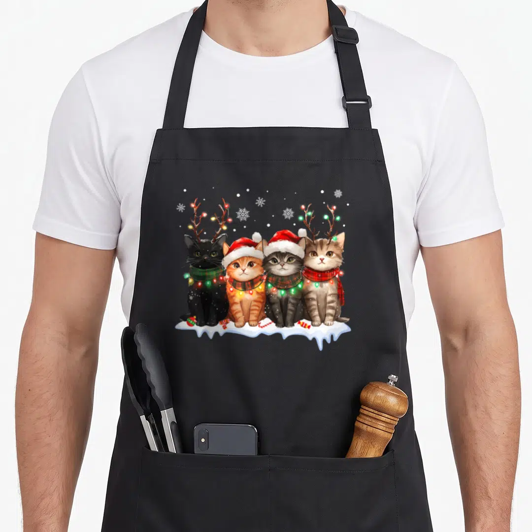 Cat reindeer apron - Four cartoon cats on snow with antlers, scarves, and colored lights — shown on a black cotton canvas apron against a white background