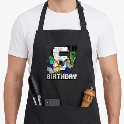 Shot birthday apron - Pixel art 6th birthday design with Steve, Alex, zombie, creeper, and wolf characters clustered — shown on a black cotton canvas apron against a white background