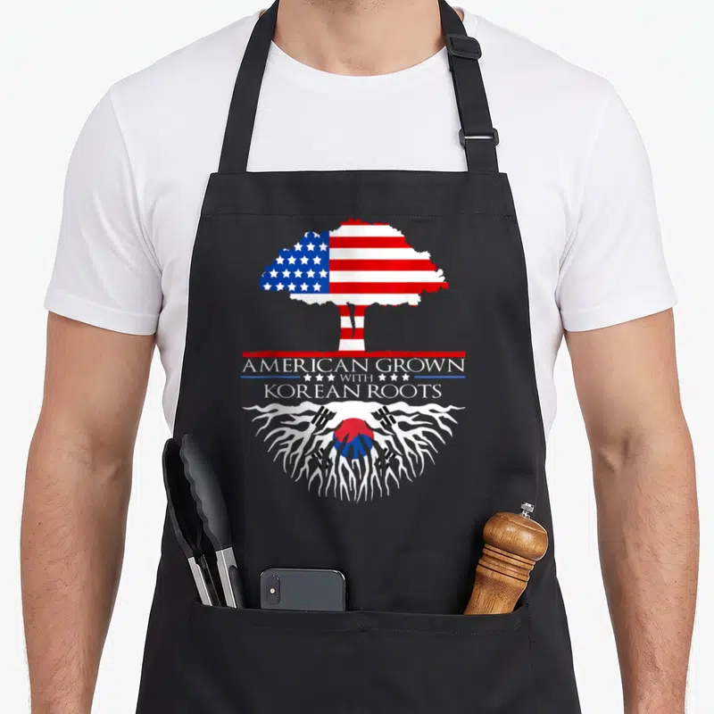 Korean asian apron - Tree with US flag canopy and Korean flag roots surrounding stacked text AMERICAN GROWN WITH KOREAN — shown on a black cotton canvas apron against a white background