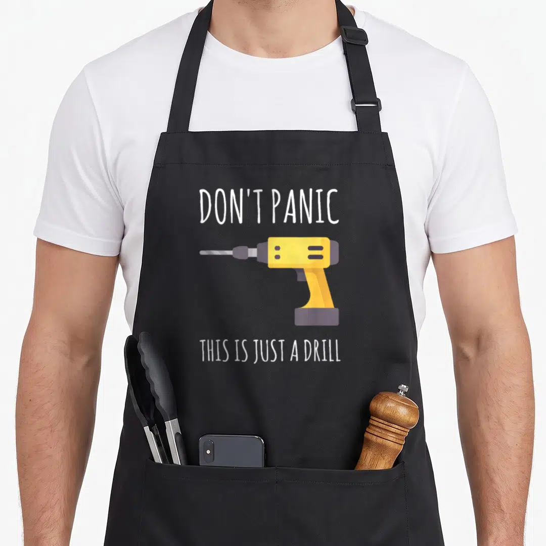 Panic handyman apron - Cartoon yellow drill between white text reading DON'T PANIC THIS IS JUST A DRILL — shown on a black cotton canvas apron against a white background