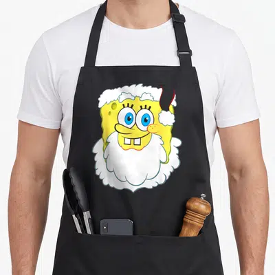 Squarepant santa apron - Yellow cartoon character face with white beard and blue eyes — shown on a black cotton canvas apron against a white background