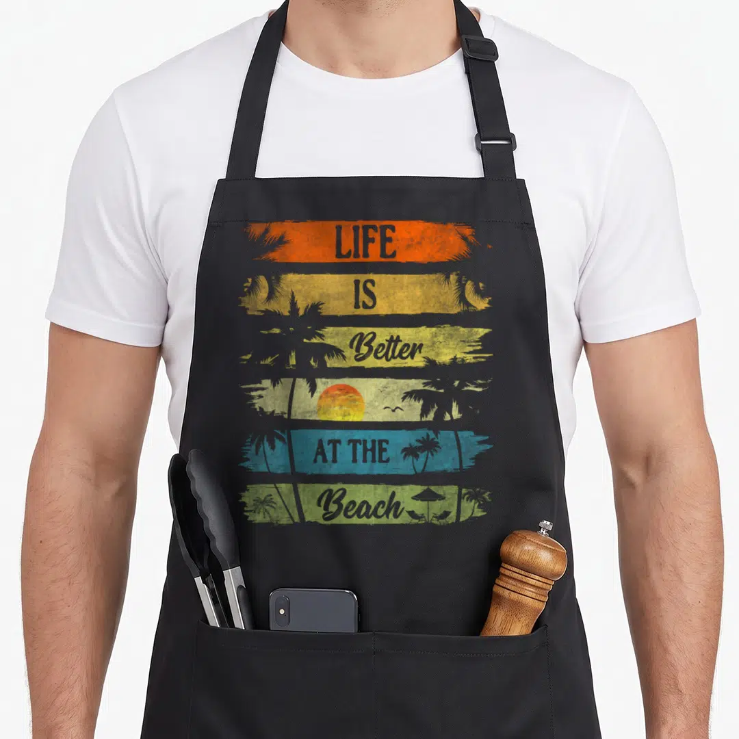 Vacation beach summer aprons - LIFE IS Better AT THE Beach text in orange yellow blue and green brushstrokes with palm — shown on a black cotton canvas apron against a white background