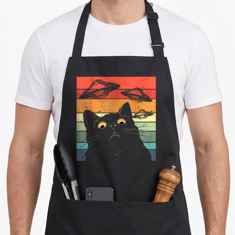 Spaceship cat apron - Grey cat head on rainbow stripes with three grey UFOs in retro art style — shown on a black cotton canvas apron against a white background