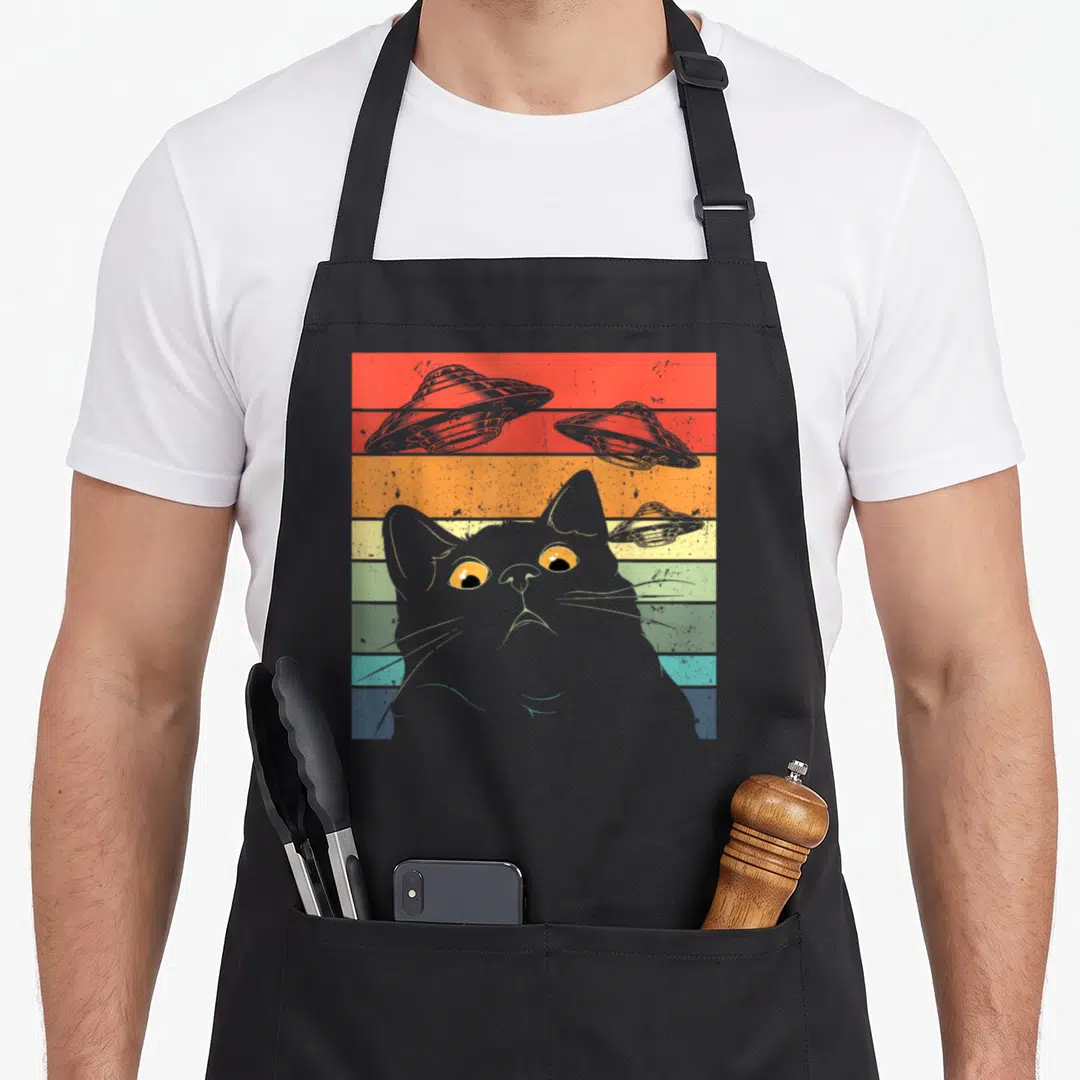 Spaceship cat apron - Grey cat head on rainbow stripes with three grey UFOs in retro art style — shown on a black cotton canvas apron against a white background