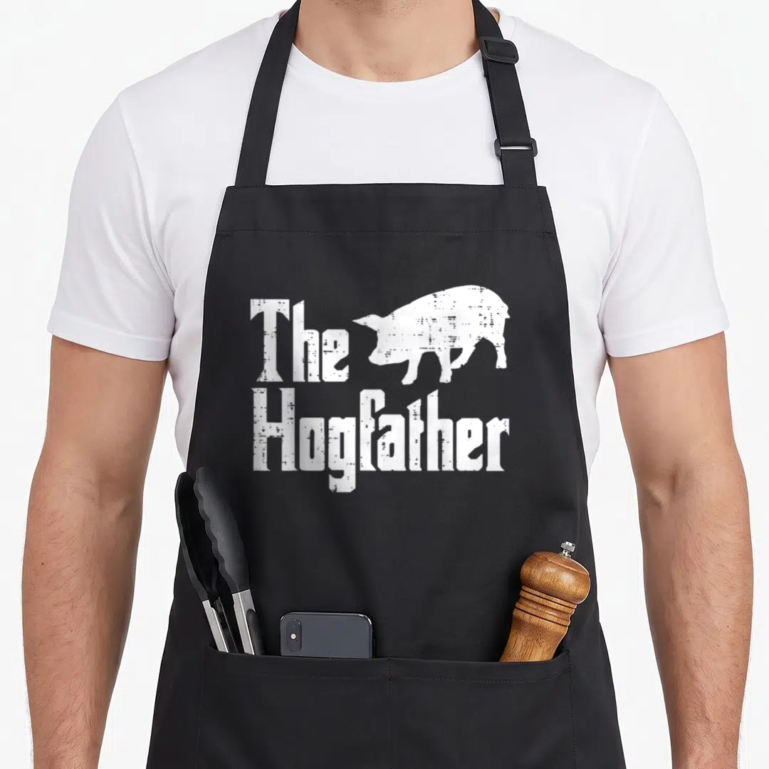 Pig father day apron - White distressed text The Hogfather with pig silhouette — shown on a black cotton canvas apron against a white background