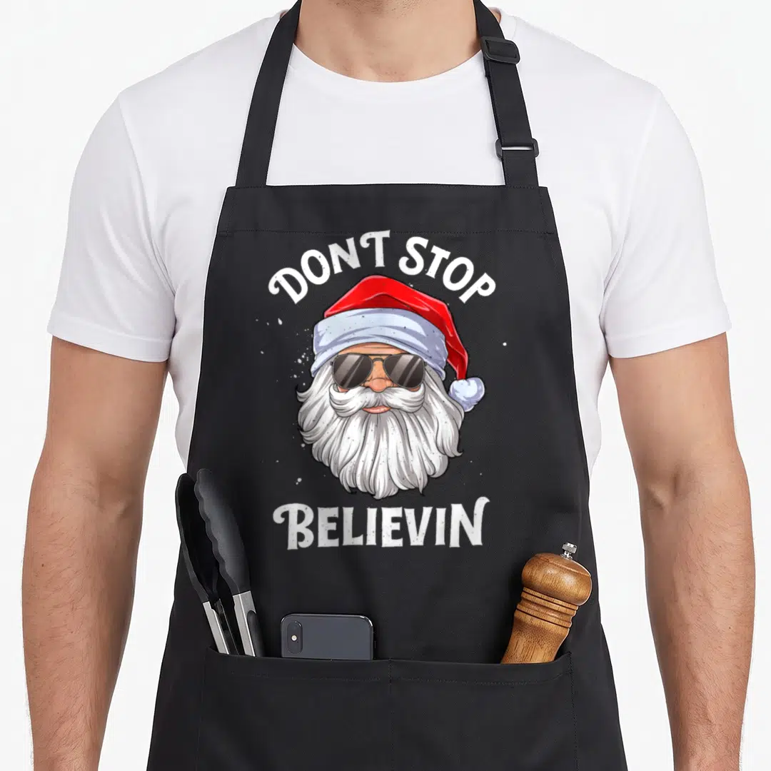 Stop santa apron - Cartoon Santa face with sunglasses and text DONT STOP BELIEVIN in red and white — shown on a black cotton canvas apron against a white background