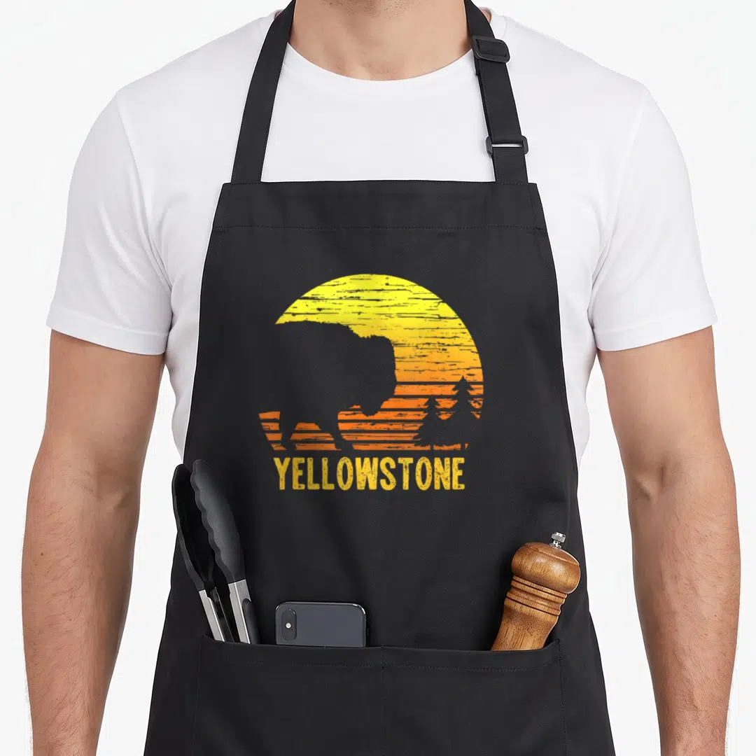 Vintage wyoming buffalo apron - Grey bison silhouette in yellow orange sunset circle with YELLOWSTONE text below — shown on a black cotton canvas apron against a white background